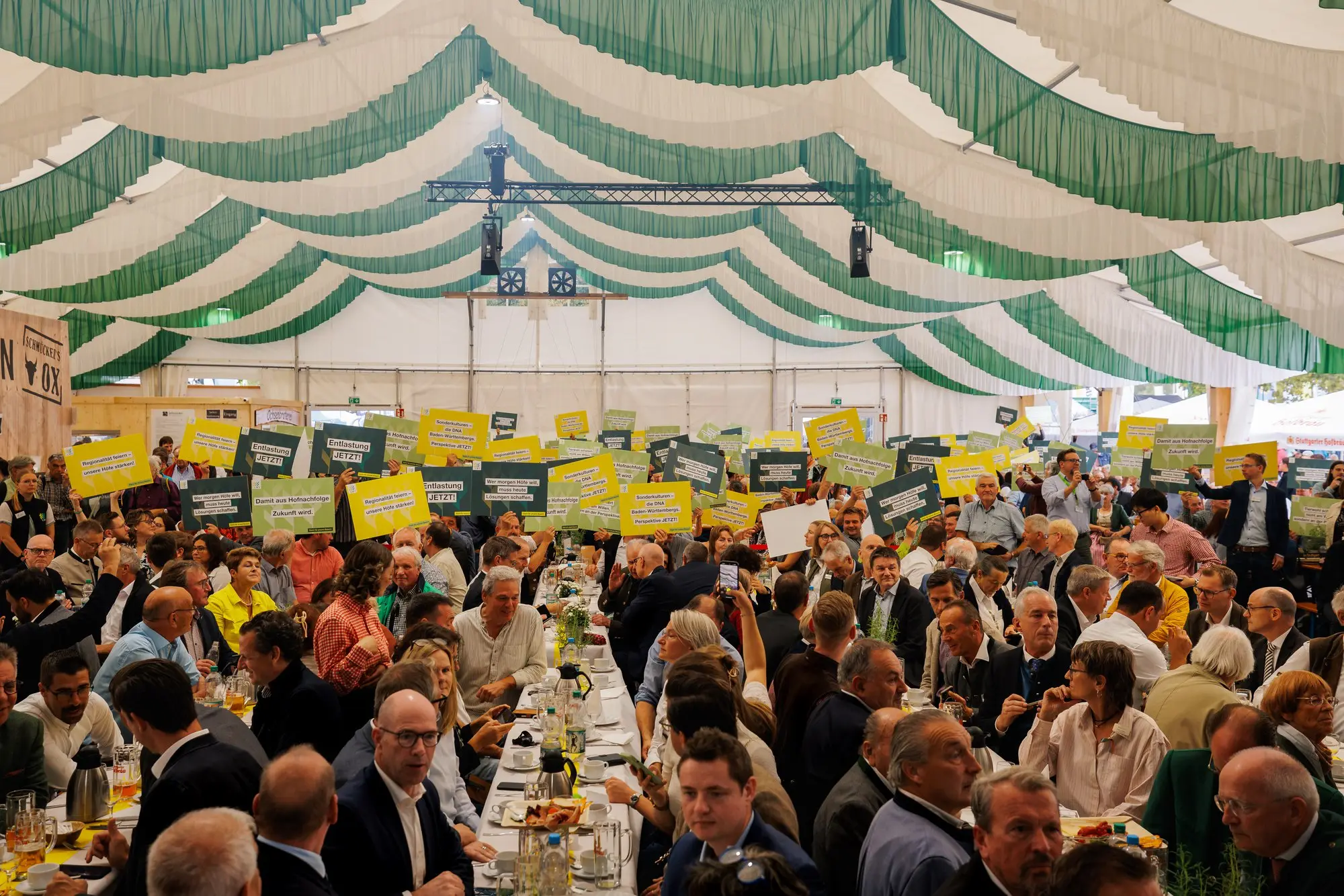Gäste im Festzelt halten gelbe und grüne Schilder mit Forderungen der Landwirtschaft hoch